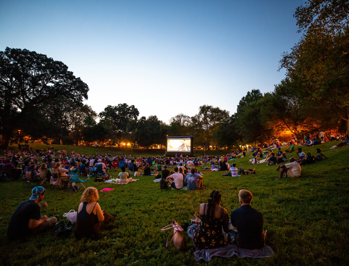 A large group of people sitting on the ground, watching a movie on a big screen during Movies at Clark Park