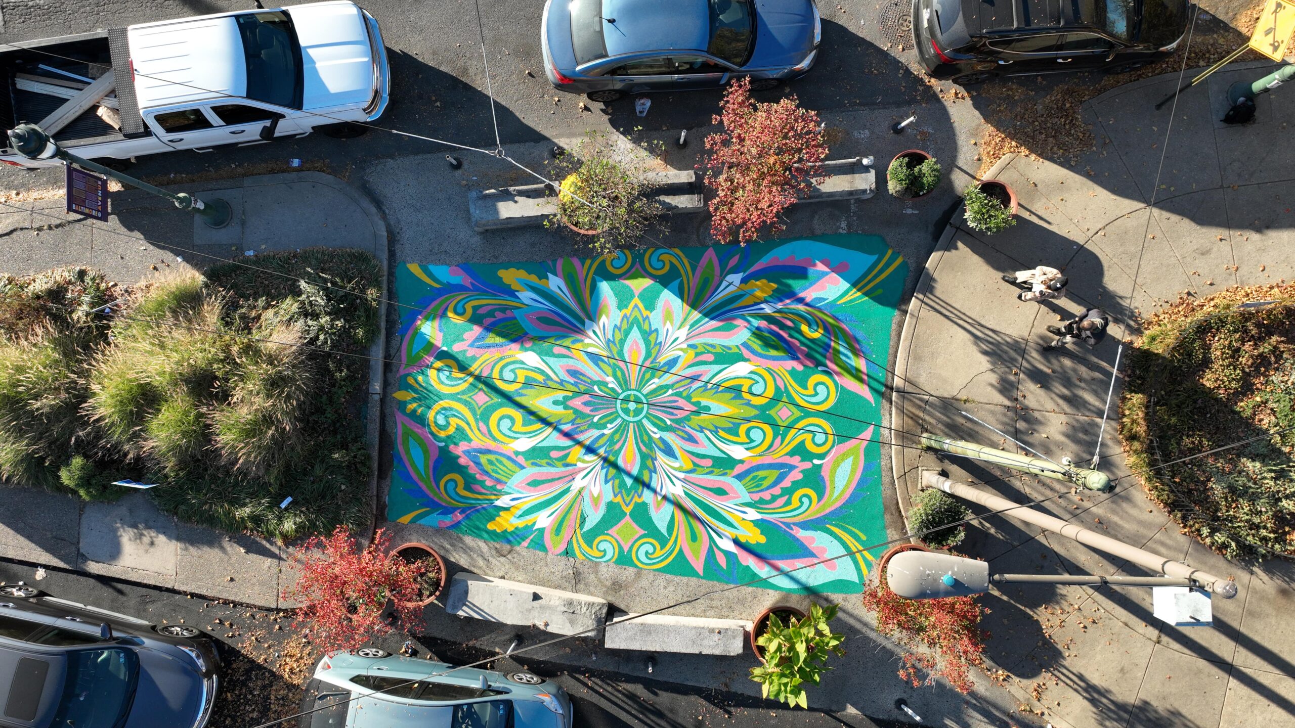 An aerial view of a colorful mural at Cedar Park Pedestrian Plaza, surrounded by trees, parked cars, and pedestrians on a sunny day.