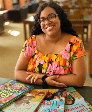 Headshot of author Alyssa Reynoso-Morris. She sits at a tble with arms crossed, wearing a colorful flowery shirt and smiling
