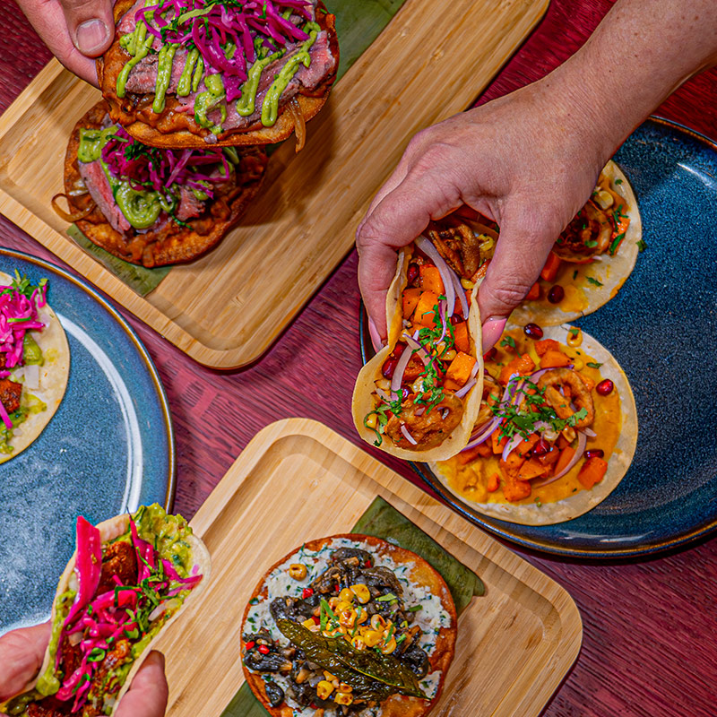 A promotional image from El Mictlan restaurant, features people sharing tacos over a colorful table