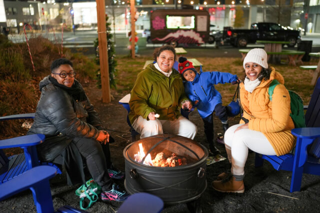 A group of people sit at an outdoor bonfire, smiling and facing the camera. They all wear winter gear.