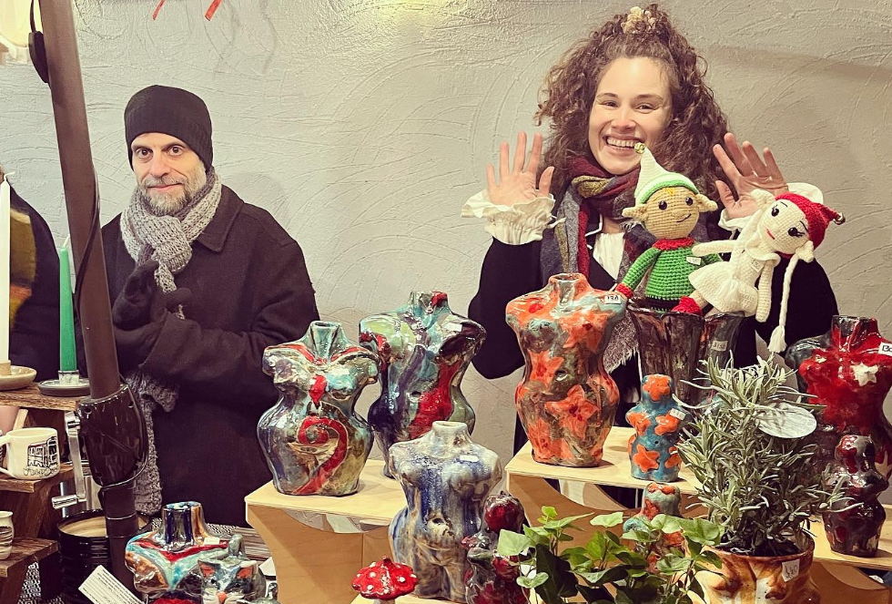 Two people at an outdoor market, smiling and waving at the camera, selling christmas gifts