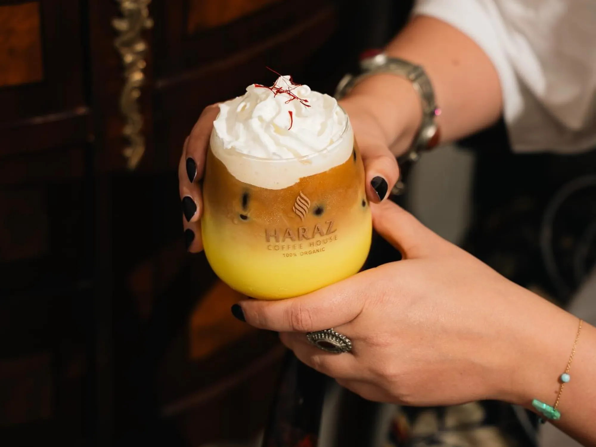 Hands holding a Haraz Coffee branded cup, the drink its yellow, topped with espresso, and topped with whipped cream