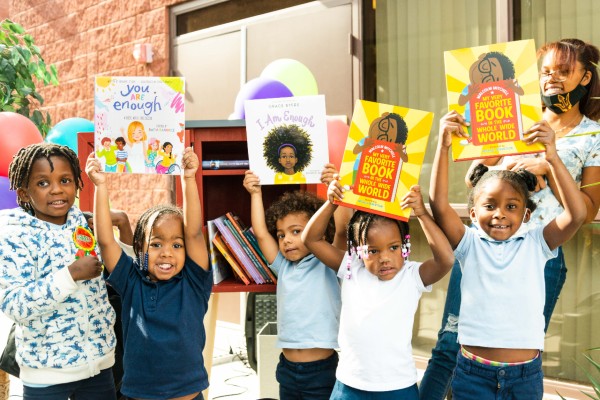 A group of children stand outside of a library, holding books and smiling