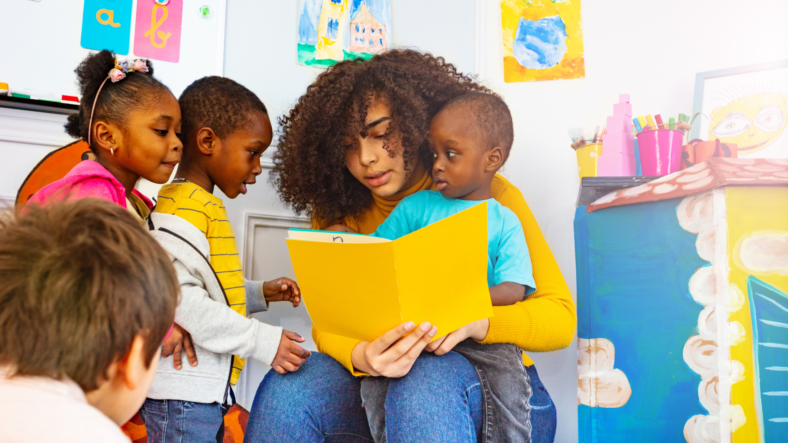 A group of kids crowd around a teacher as she sits, and reads them a story.