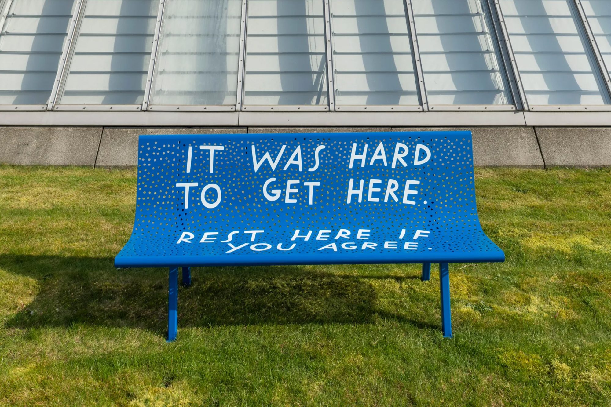 Blue outdoor bench on a grassy lawn with white text reading “It was hard to get here. Rest here if you agree,” set in front of a modern glass building.