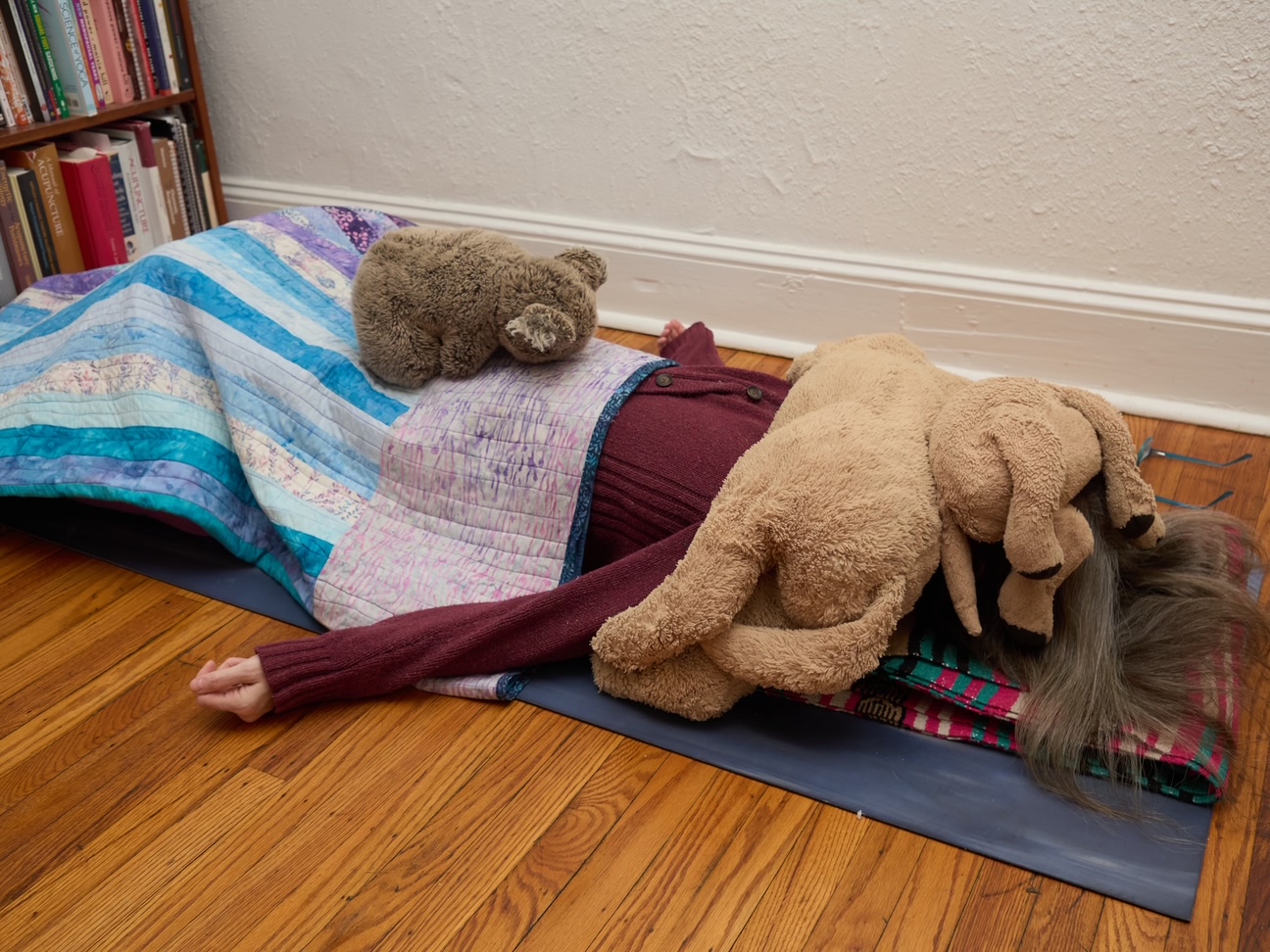 A person lays on the ground surrounded by blankets and stuffed animals during a yoga session