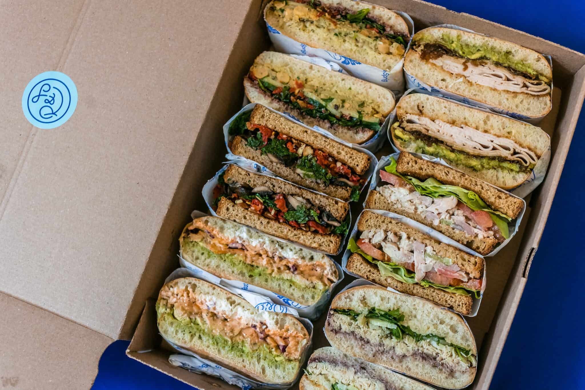 Several sandwiches packed with meat, veggies, and sauce are displayed together in a cardboard box.