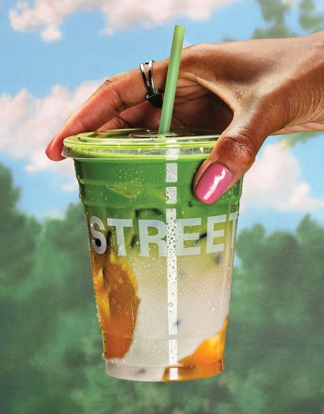 A Black woman's hand is holding a plastic cup with a green, white, and orange beverage inside of it. Her nails are painted pink. The background is a blue sky with clouds and green trees,